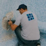 Operario con pistolete retirando gresite de la pared de una piscina vacía en proceso de reparación.