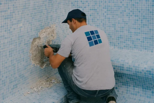 Operario con pistolete retirando gresite de la pared de una piscina vacía en proceso de reparación.